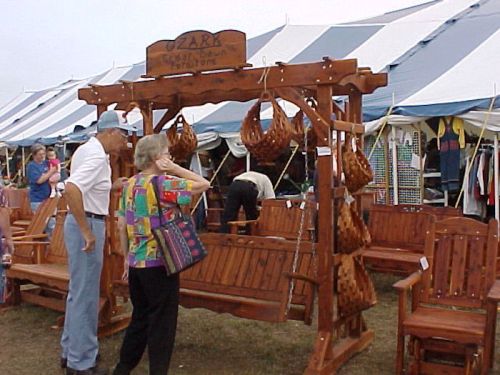 A wooden swing set with hanging baskets, displayed at a fair, with people browsing nearby under a large tent.