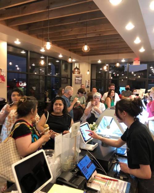 A busy café scene with a crowd of customers at the counter, interacting with staff and looking at the menu.