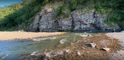 A serene river flows past rocky cliffs, surrounded by lush greenery and a person standing by the water's edge.