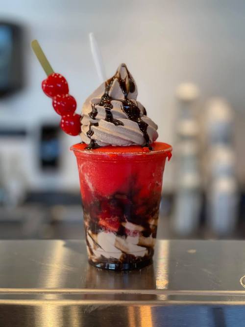 A colorful dessert cup with chocolate soft serve, cherry topping, and a red syrup base, garnished with cherries.