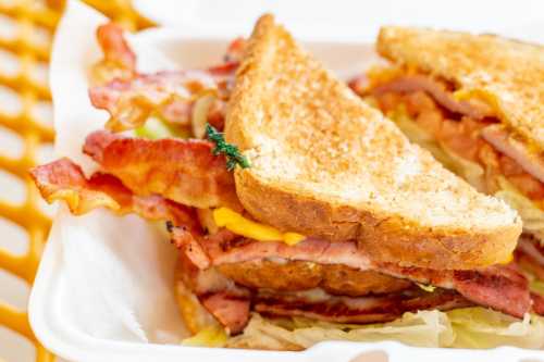 A close-up of a toasted sandwich filled with bacon, ham, cheese, and lettuce, served in a takeout container.