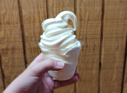A hand holding a cup of soft-serve vanilla ice cream with a swirl on top, against a wooden background.