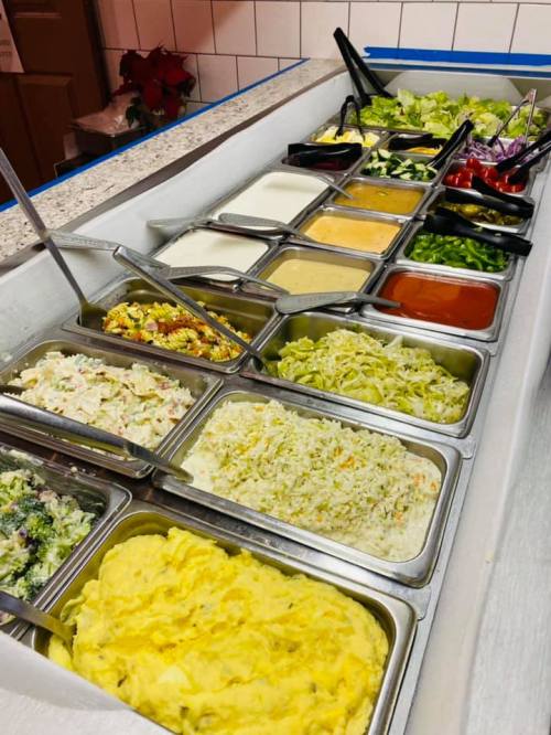 A variety of salad and condiment options in metal trays, including greens, dressings, and toppings on a countertop.