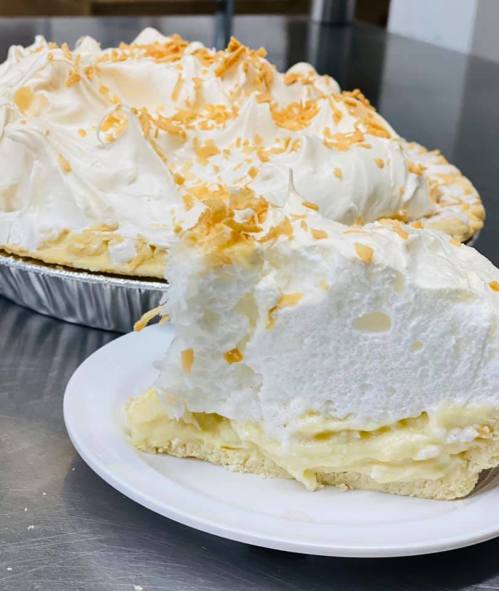 A slice of coconut cream pie on a plate, with a whole pie in the background, topped with whipped cream and toasted coconut.