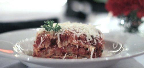 A plate of lasagna topped with grated cheese and a sprig of parsley, served in a restaurant setting.