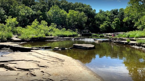 A serene natural landscape featuring a calm stream, rocky banks, and lush greenery under a clear blue sky.