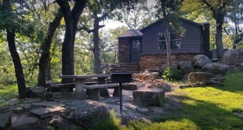 A cozy cabin surrounded by trees, with a stone patio and picnic table in a natural setting.