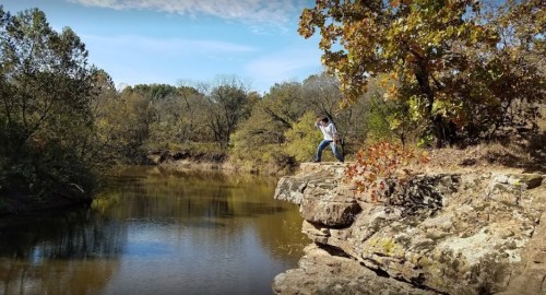 A person stands on a rocky ledge by a calm river, surrounded by trees and a clear blue sky.