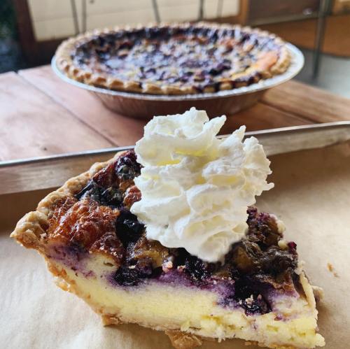 A slice of blueberry pie topped with whipped cream, with a whole pie in the background on a wooden table.