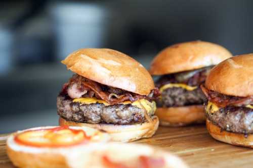 Three delicious burgers with bacon and cheese on toasted buns, with a slice of tomato and ketchup on the side.