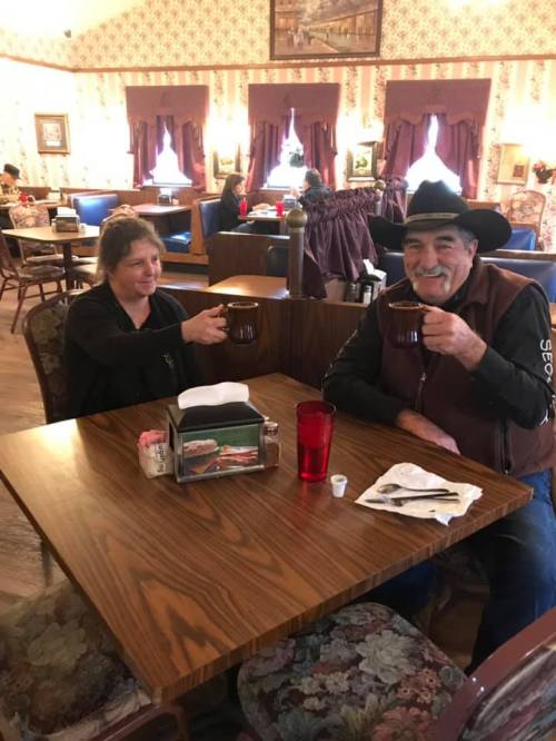 Two people sitting at a table in a cozy restaurant, smiling and holding large mugs of coffee.