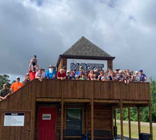 A large group of people poses on the balcony of a wooden building labeled "The Maze" under a cloudy sky.