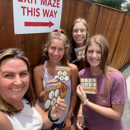 A group of four young women smiling and holding game cards in front of a maze exit sign.