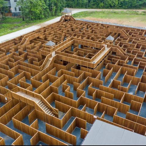 Aerial view of a large wooden maze with multiple pathways and two elevated platforms. Surrounded by trees.