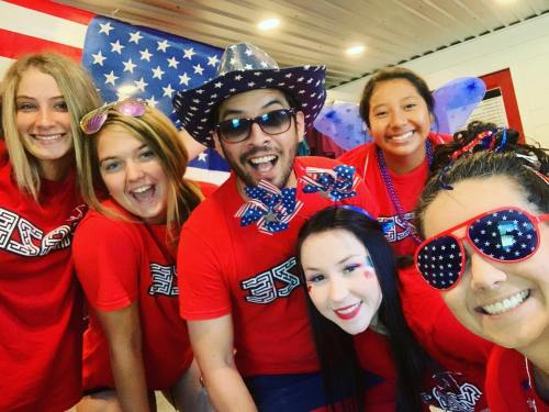 A group of six people in red shirts, festive accessories, and patriotic decorations, smiling and celebrating together.