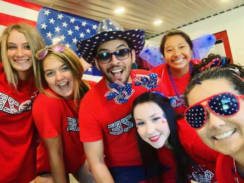 A group of six people in red shirts, festive accessories, and patriotic decorations, smiling and celebrating together.