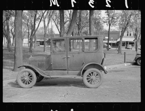 A vintage, dusty car parked on a tree-lined street, with buildings visible in the background.
