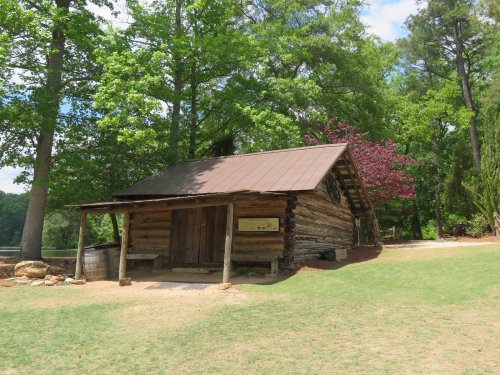 A rustic log cabin surrounded by green trees and grass, with a metal roof and a dirt path leading to it.