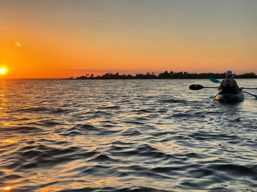 A person kayaking on calm water at sunset, with vibrant orange and yellow hues in the sky.