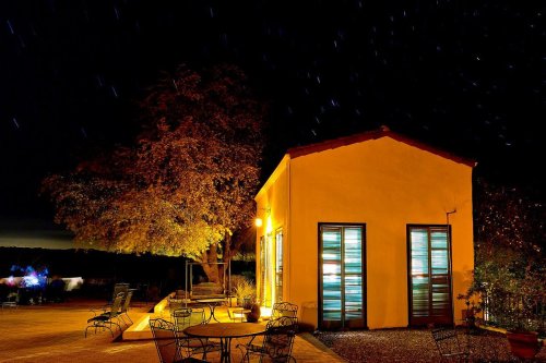 A cozy yellow building at night, surrounded by trees and outdoor seating, with stars visible in the dark sky.