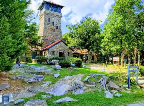 A stone tower and building surrounded by greenery and rocks under a blue sky with scattered clouds.