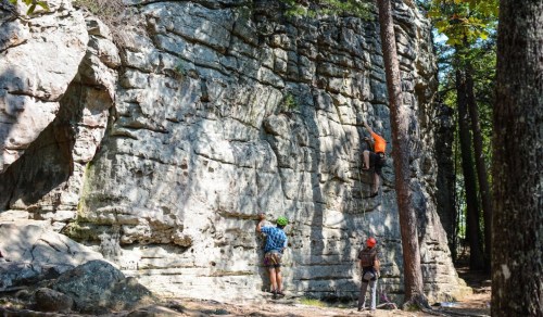 Two climbers scale a rocky wall in a forest, while another person observes from the ground. Trees surround the area.