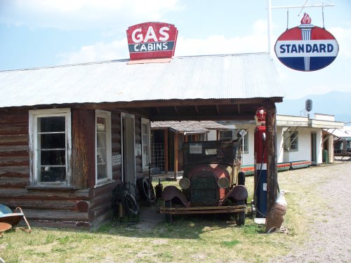 A vintage gas station with a classic car and cabins, featuring a "Gas" sign and a "Standard" gas logo.