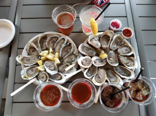 Two plates of oysters with lemon wedges, surrounded by drinks and dipping sauces on a table.