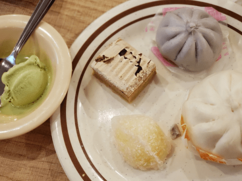 A plate with various desserts: a green ice cream scoop, a purple dumpling, a white dumpling, and two sweet pastries.