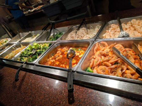 A buffet display featuring various dishes, including shrimp, vegetables, noodles, and sauces in metal trays.