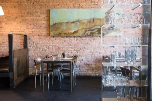 A modern restaurant interior featuring a wooden table, chairs, and a large abstract painting on a brick wall.