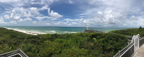 A panoramic view of a beach with green vegetation, a wooden walkway, and a cloudy blue sky.