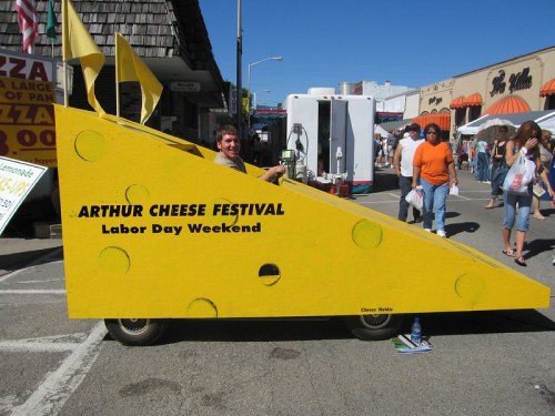 A person sits in a large, yellow cheese-shaped vehicle at the Arthur Cheese Festival, with festival-goers in the background.