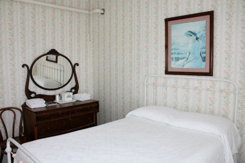 A simple bedroom with a white bed, vintage dresser, mirror, and floral wallpaper. A framed painting hangs on the wall.