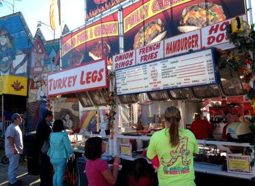 A food stand at a fair featuring turkey legs, onion rings, fries, hamburgers, and hot dogs with a colorful backdrop.
