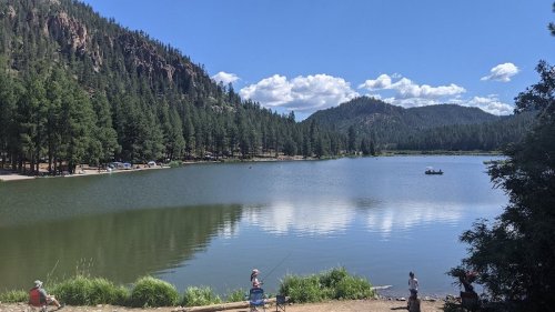 A serene lake surrounded by trees and mountains, with people fishing and enjoying the outdoors on a sunny day.