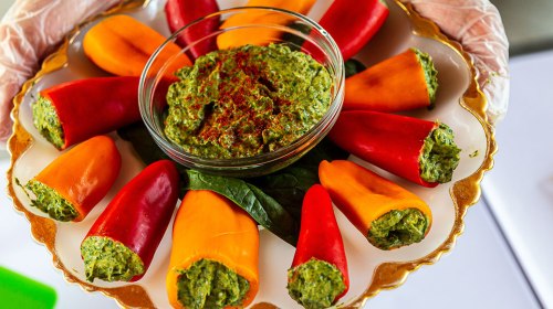 A platter of stuffed mini peppers in red and yellow, surrounded by a bowl of green dip, garnished with spices.