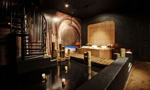 A luxurious spa room with a spiral staircase, a sunken tub, and candles, featuring an elegant archway mural.