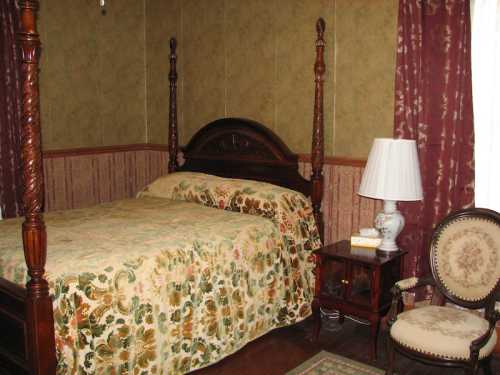 A cozy bedroom featuring a four-poster bed, floral bedding, a lamp, and a vintage chair against patterned wallpaper.