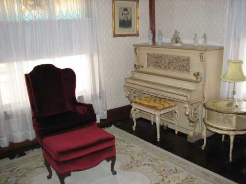 A vintage room featuring a red velvet chair, an ornate piano, and a decorative lamp, with soft natural light.