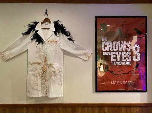 A stained white lab coat with black feathers hangs next to a poster for "The Crows Have Eyes 3: The Crowening."