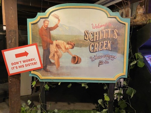 Sign for "Schitt's Creek" featuring a humorous illustration and a warning sign that says, "Don't worry, it's his sister!"