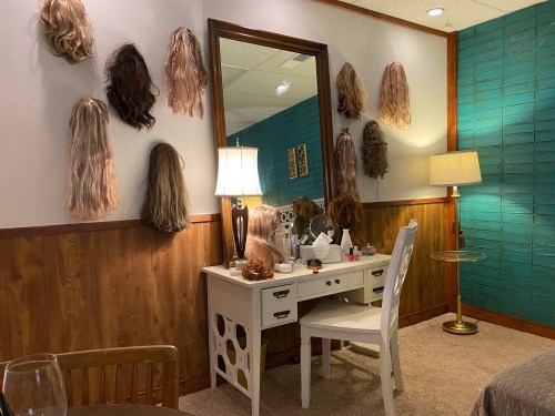 A room with a mirror, a vanity table, and various wigs displayed on the wall, featuring a lamp and a chair.