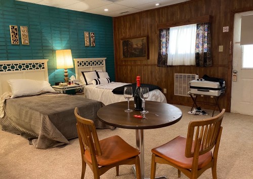 Cozy room with two beds, a table for two, a bottle of wine, and a window with curtains. Warm, inviting decor.