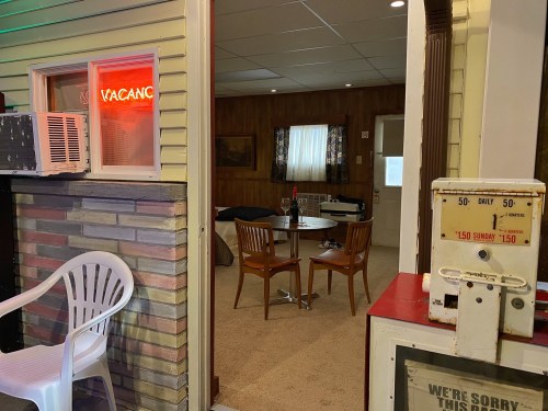 A cozy room with a "VACANCY" sign, featuring a table, chairs, and vintage decor, seen from an outdoor perspective.