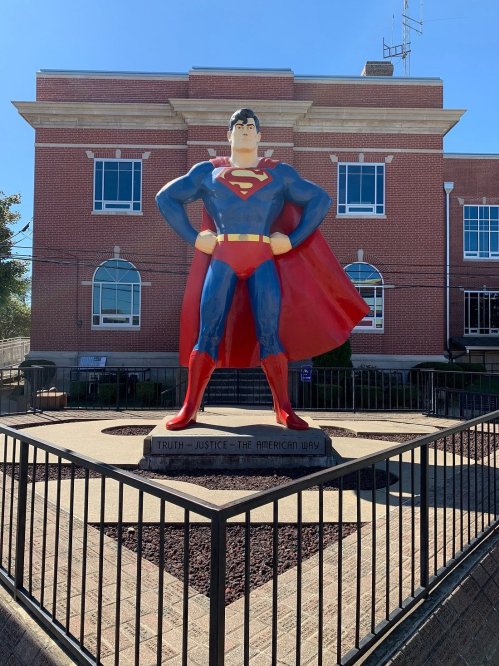 A large Superman statue stands in front of a brick building, with a sign reading "Truth - Justice - The American Way."