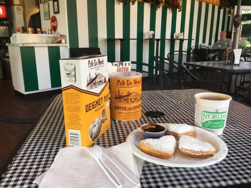 A table with beignets, powdered sugar, a cup of coffee, and boxes from Café Du Monde in a striped café setting.