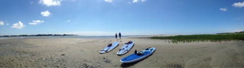 Two people stand on a sandy beach near three paddleboards under a clear blue sky.