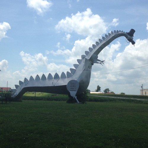 A large metal dinosaur sculpture with a long neck and spiked back, set against a blue sky and green grass.