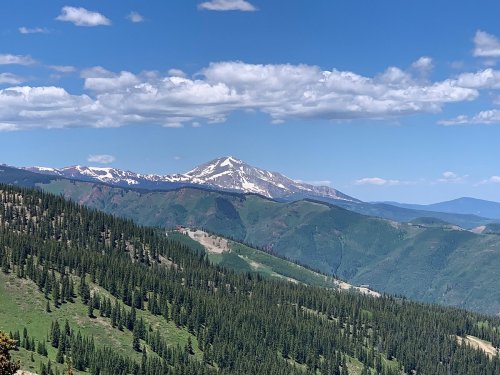 A scenic view of a snow-capped mountain surrounded by lush green forests and blue skies with scattered clouds.
